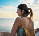 Young adult woman with cochlear implant walking confidently by the sea during a summer vacation. Her hair is tied in a ponytail, revealing the implant and symbolizing independence, adventure and inclusivity.