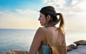 Young adult woman with cochlear implant walking confidently by the sea during a summer vacation. Her hair is tied in a ponytail, revealing the implant and symbolizing independence, adventure and inclusivity.