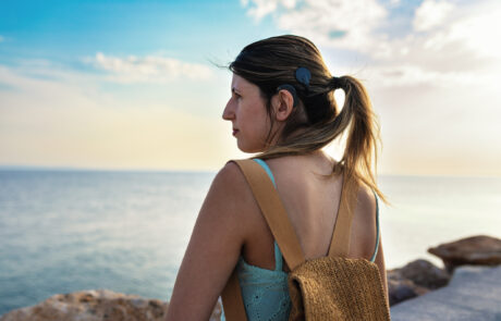 Young adult woman with cochlear implant walking confidently by the sea during a summer vacation. Her hair is tied in a ponytail, revealing the implant and symbolizing independence, adventure and inclusivity.