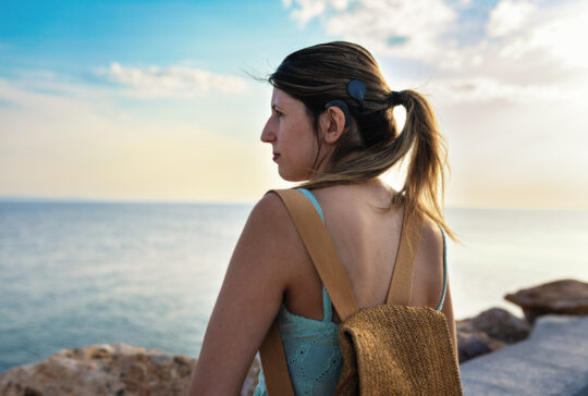 Young adult woman with cochlear implant walking confidently by the sea during a summer vacation. Her hair is tied in a ponytail, revealing the implant and symbolizing independence, adventure and inclusivity.