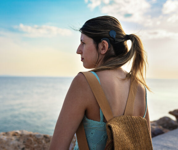 Young adult woman with cochlear implant walking confidently by the sea during a summer vacation. Her hair is tied in a ponytail, revealing the implant and symbolizing independence, adventure and inclusivity.