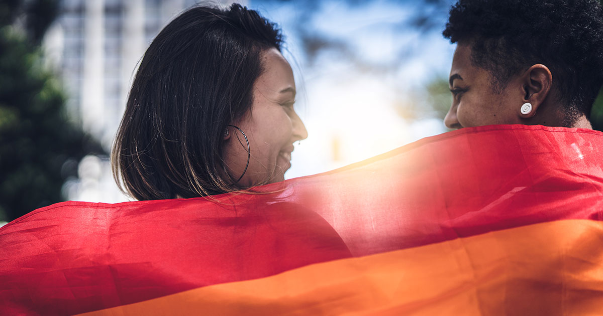 Two people smiling and holding up a rainbow Pride flag behind their shoulders.jpg