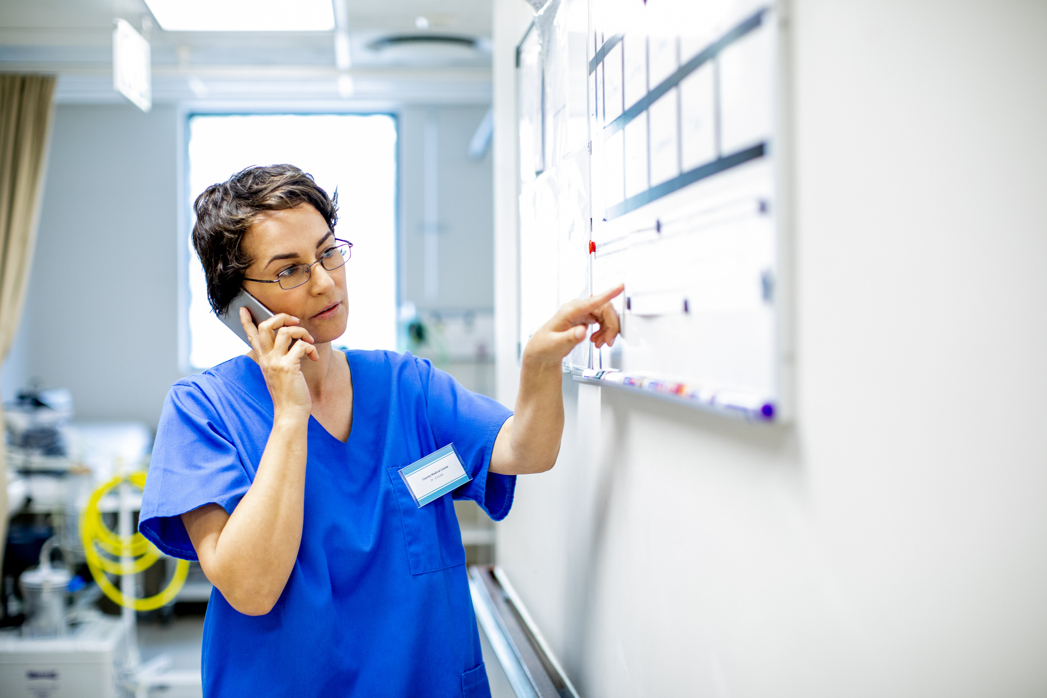 Young female doctor standing in the corridor of a modern hospital reading a scheduling whiteboard and talking on a cellphone