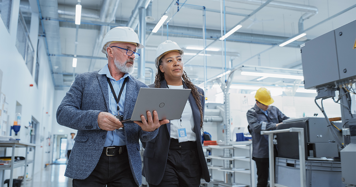 Two Professional Heavy Industry Engineers Wearing Hard Hats at Factory. Walking and Discussing Industrial Machine Facility, Working on Laptop. African American Manager and Technician at Work