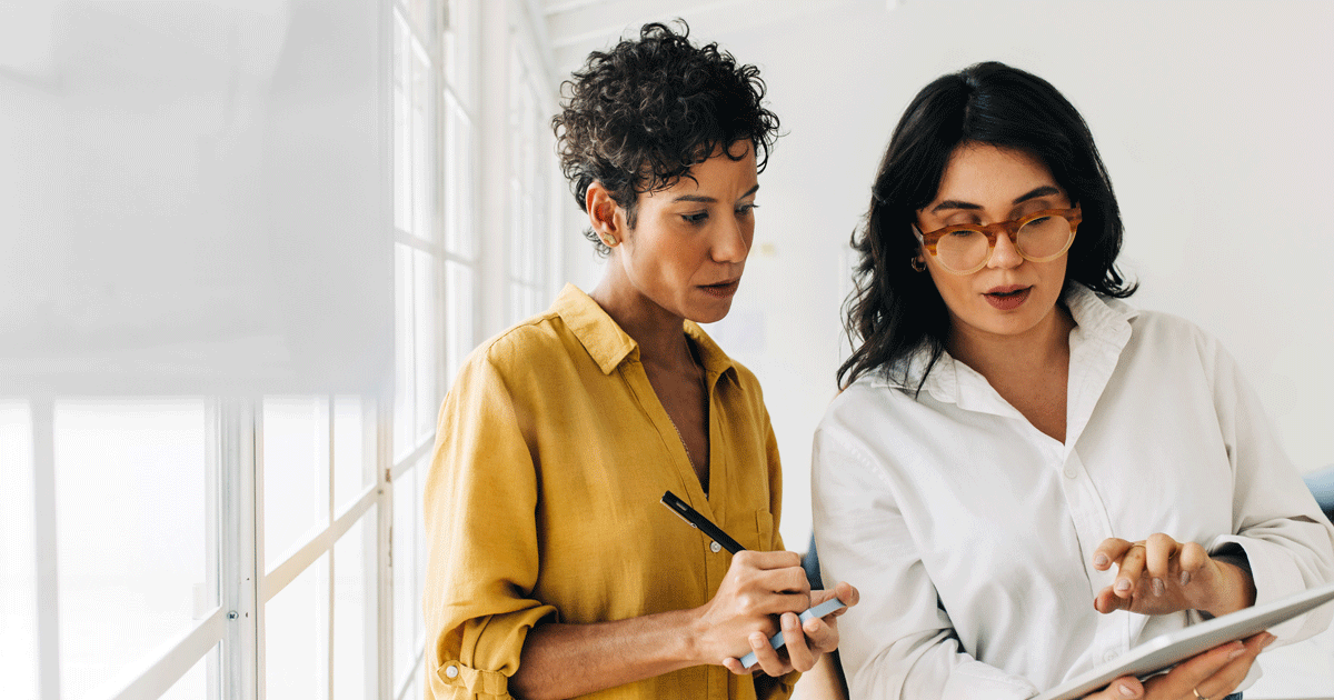 Two business women having a discussion, they're standing in an office and using a tablet