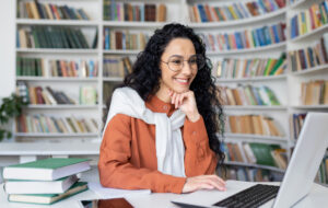 Female student with laptop reading online video course, Latin American woman smiling and satisfied with independent online learning sitting inside university campus in library - stock photo