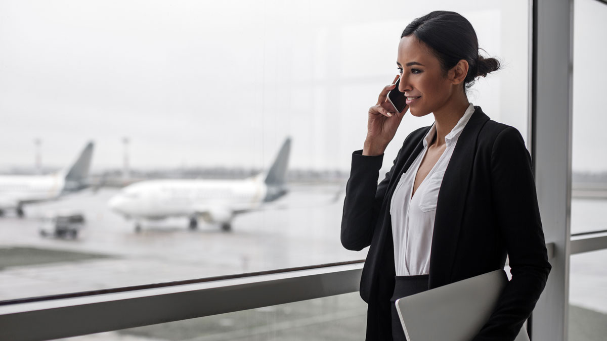 Work everywhere. Side view of young smiling woman is standing at airport lounge with laptop and looking through the window while enjoying conversation on mobile. Copy space and aircrafts in background