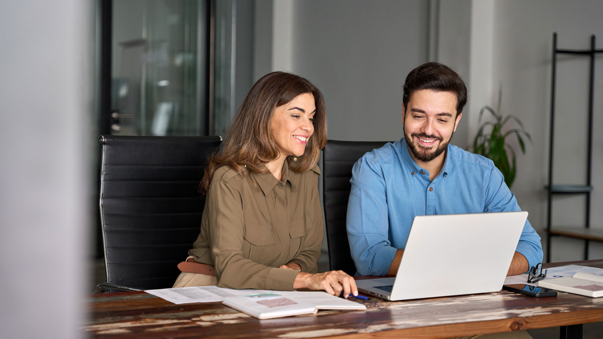 Two busy happy professional business people working in office with laptop computer. Middle aged female executive manager talking to male colleague having conversation sitting at workplace