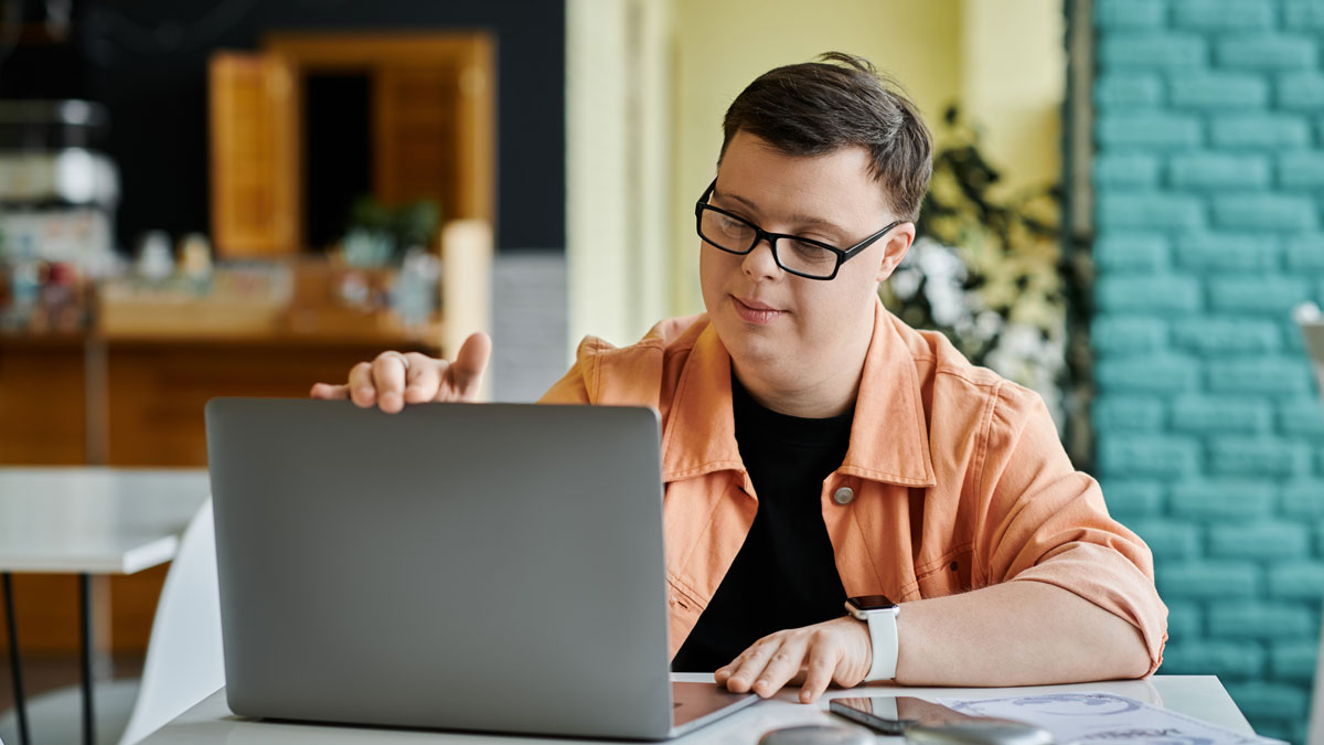 A young man with Down Syndrome is working on his laptop in a cafe, focused on his task
