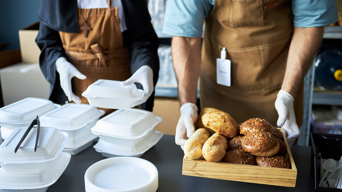 Restaurant staff preparing food orders with hands seen organizing takeaway boxes and serving freshly baked bread on a tray in the kitchen. Workers wearing aprons and gloves