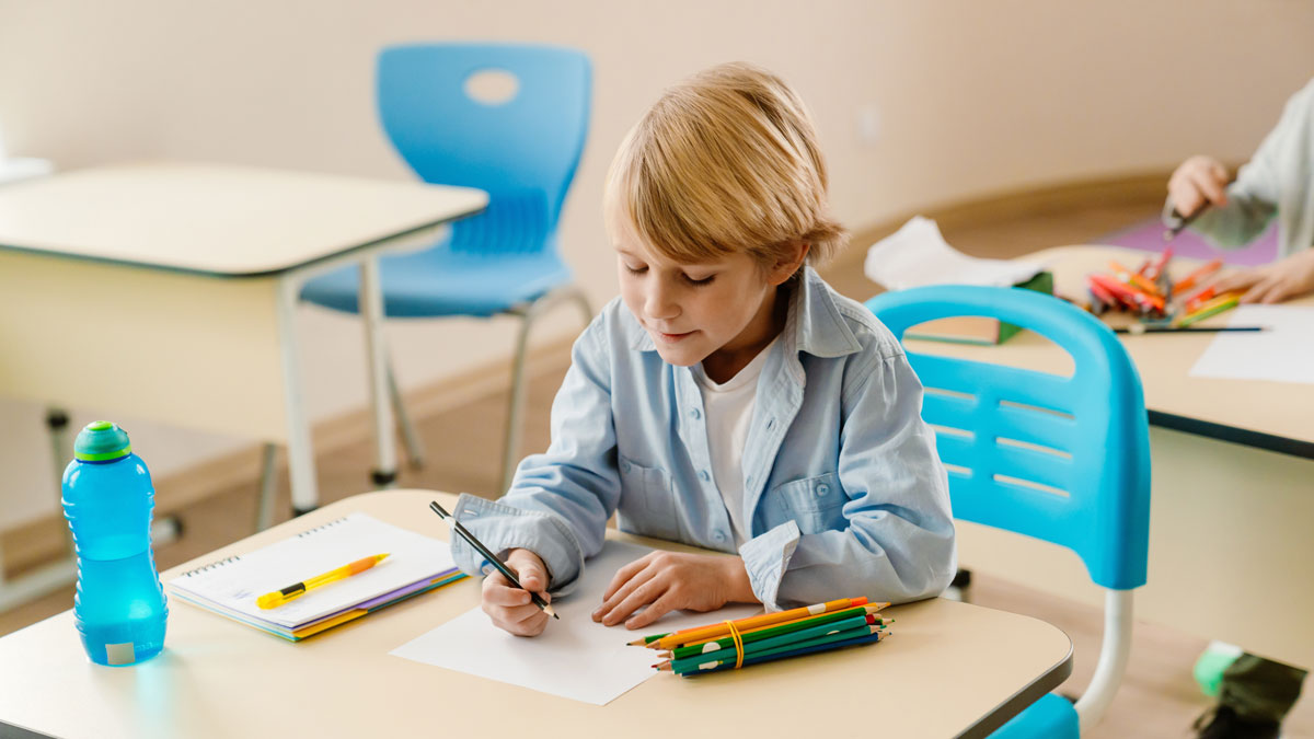 A school-aged White boy sits at a desk in a classroom drawing with colored pencils. The child is focused on his schoolwork during a creative activity in a modern learning environment