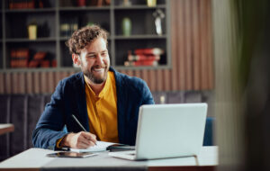 Young smiling bearded Caucasian blogger dressed smart casual writing notes in agenda and looking at laptop while sitting in cafeteria.