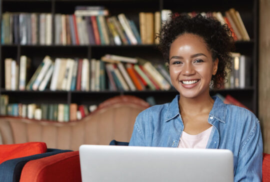 Cheerful female writer with attractive appearance wearing jean clothes sitting at sofa against shelves with books, working on her new article while using laptop computer and free wi-fi connection