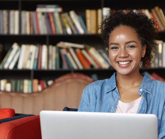 Cheerful female writer with attractive appearance wearing jean clothes sitting at sofa against shelves with books, working on her new article while using laptop computer and free wi-fi connection