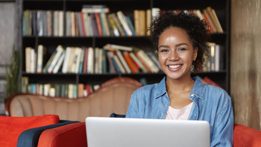 Cheerful female writer with attractive appearance wearing jean clothes sitting at sofa against shelves with books, working on her new article while using laptop computer and free wi-fi connection