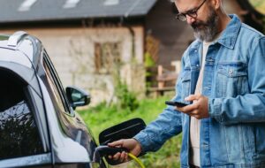 Man charging electric car in front of his house, plugging the charger into the charging port. House with solar panel system on roof behind him