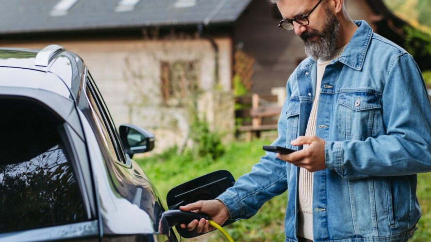 Man charging electric car in front of his house, plugging the charger into the charging port. House with solar panel system on roof behind him