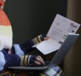 Woman In Gloves With Laptop And Bill Trying To Keep Warm By Radiator During Cost Of Living Energy Crisis