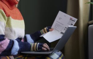 Woman In Gloves With Laptop And Bill Trying To Keep Warm By Radiator During Cost Of Living Energy Crisis