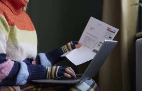 Woman In Gloves With Laptop And Bill Trying To Keep Warm By Radiator During Cost Of Living Energy Crisis