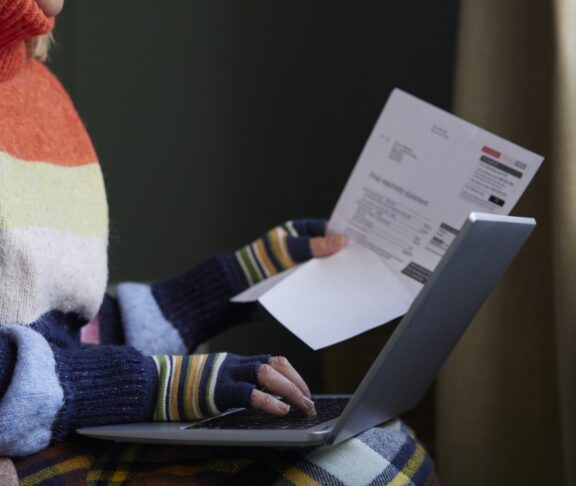 Woman In Gloves With Laptop And Bill Trying To Keep Warm By Radiator During Cost Of Living Energy Crisis