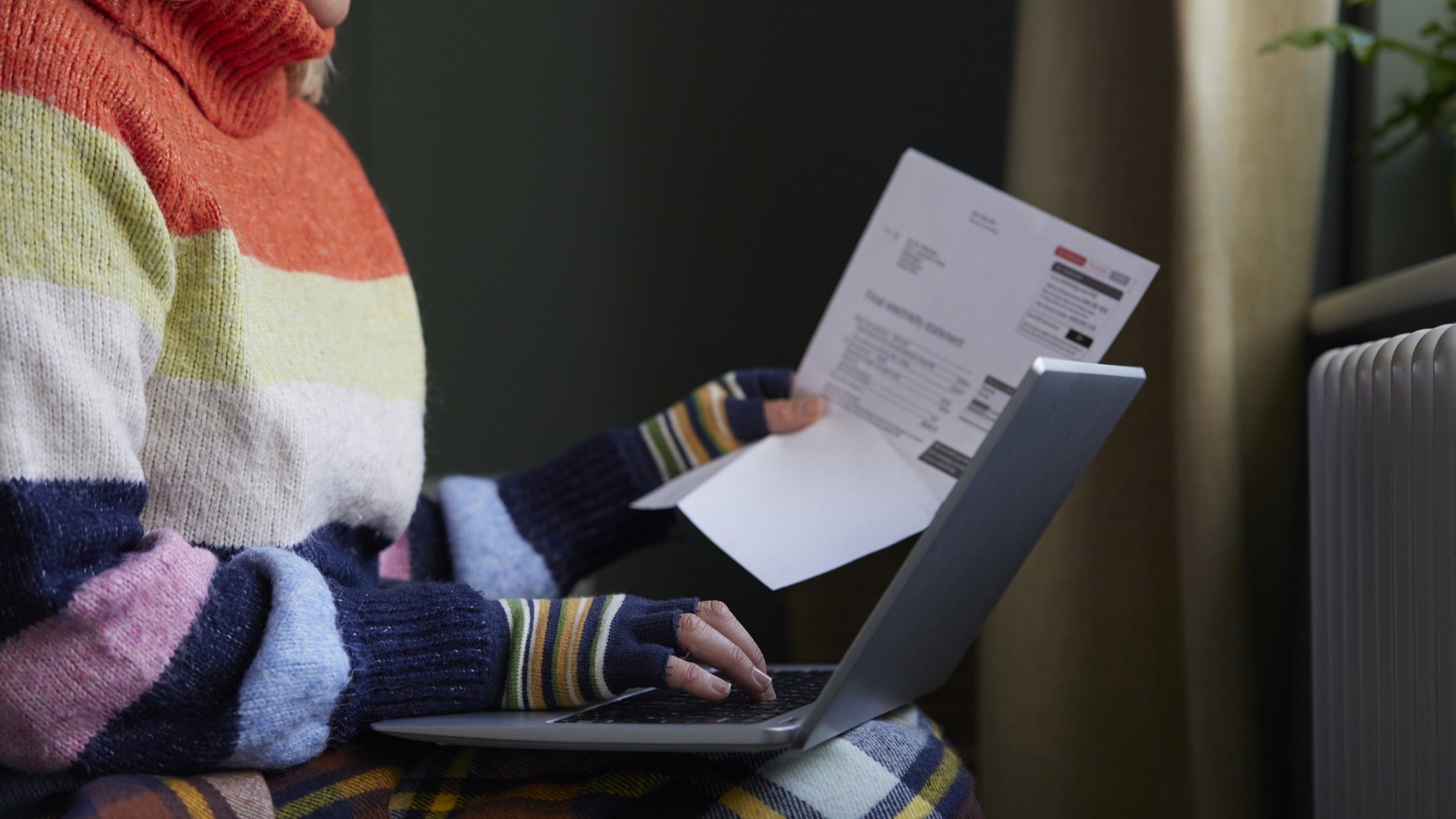 Woman In Gloves With Laptop And Bill Trying To Keep Warm By Radiator During Cost Of Living Energy Crisis