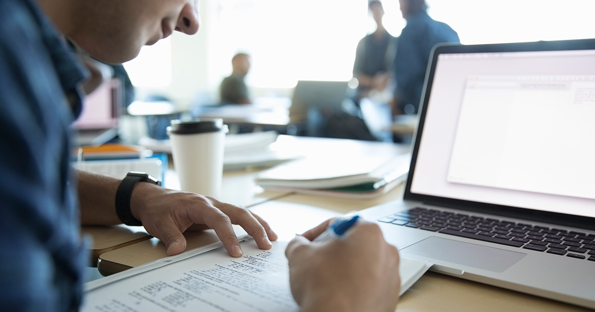 Photograph of someone writing on a notepad in front of a laptop screen