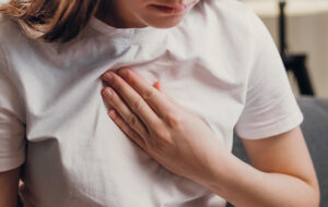 Young unhealthy female sit on couch having difficulty breathing pain of heart, touches his chest with hand