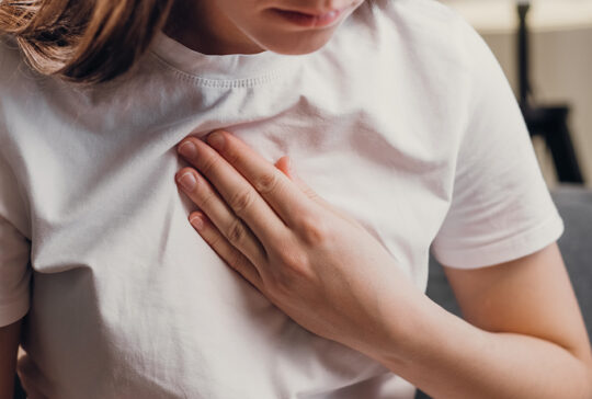 Young unhealthy female sit on couch having difficulty breathing pain of heart, touches his chest with hand