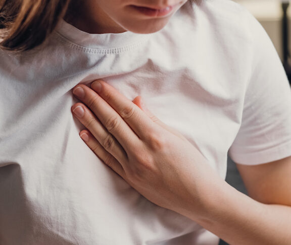 Young unhealthy female sit on couch having difficulty breathing pain of heart, touches his chest with hand