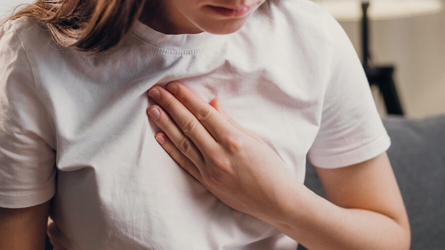 Young unhealthy female sit on couch having difficulty breathing pain of heart, touches his chest with hand
