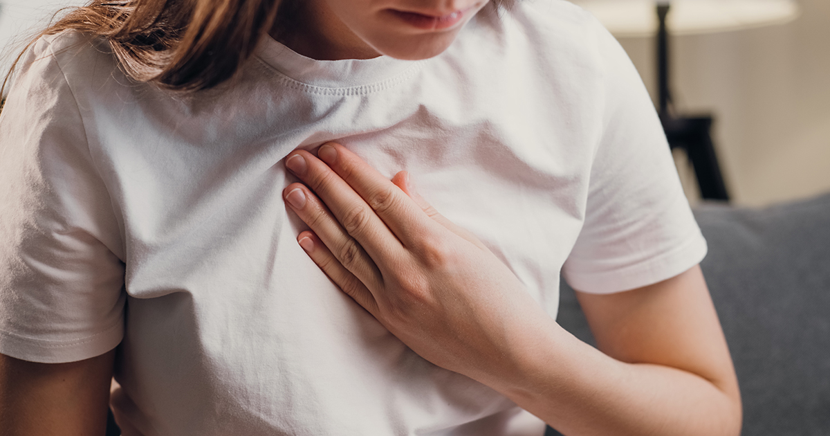 Young unhealthy female sit on couch having difficulty breathing pain of heart, touches his chest with hand
