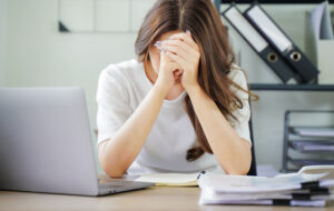 A woman is sitting at a desk with a laptop and a stack of papers