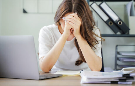 A woman is sitting at a desk with a laptop and a stack of papers