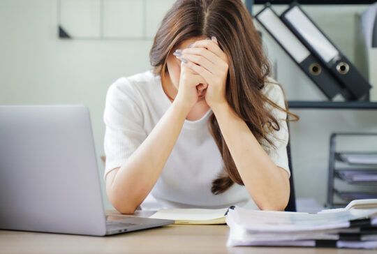 A woman is sitting at a desk with a laptop and a stack of papers