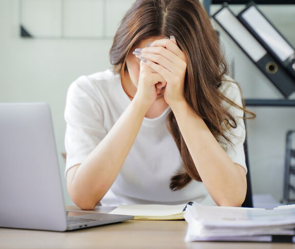 A woman is sitting at a desk with a laptop and a stack of papers