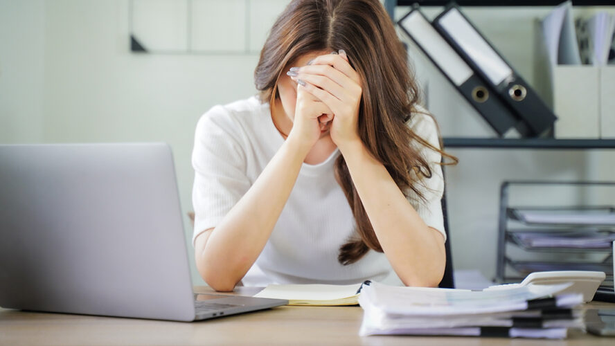 A woman is sitting at a desk with a laptop and a stack of papers