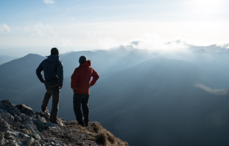 Zwei Männer stehen mit Trekking-Stöcken am Kliff und schauen auf Sonnenuntergangsstrahlen über die Wolken. Erfolgreiches Gipfelkonzept-Bild.