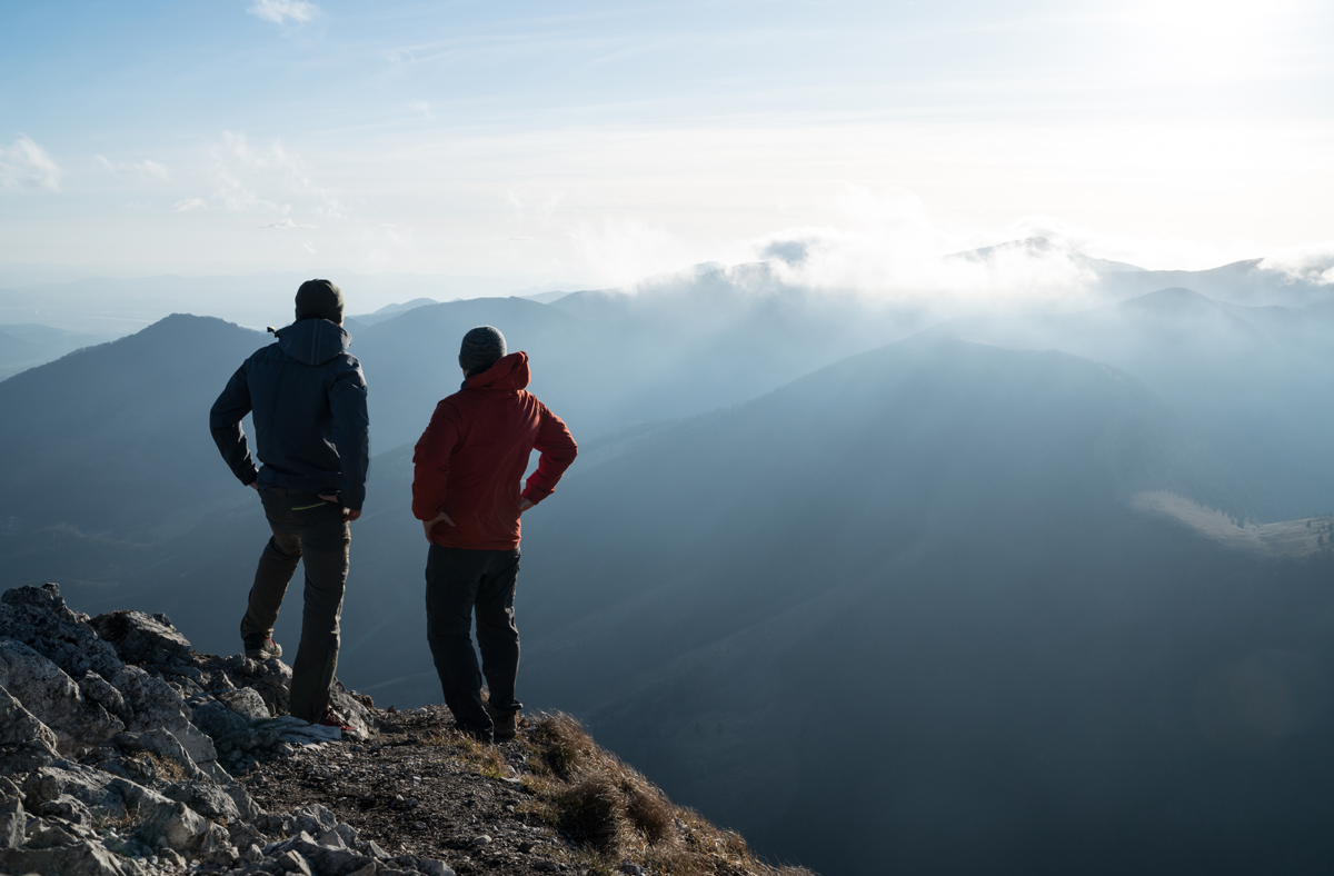 Zwei Männer stehen mit Trekking-Stöcken am Kliff und schauen auf Sonnenuntergangsstrahlen über die Wolken. Erfolgreiches Gipfelkonzept-Bild.