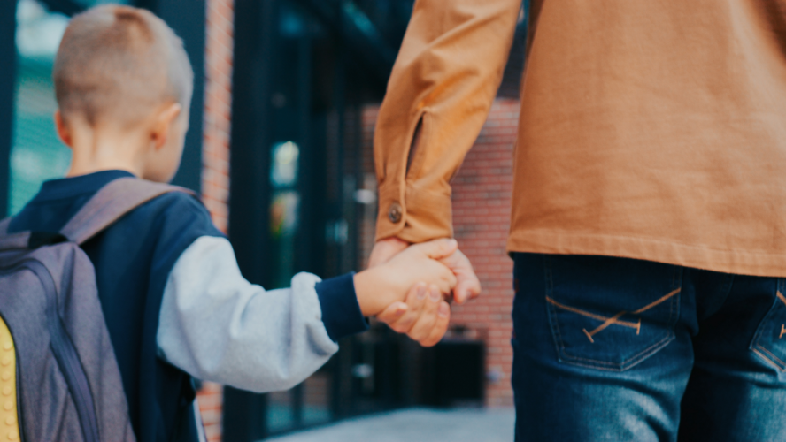 Blick von hinten auf den kleinen kaukasischen Jungen, der mit seinem fürsorglichen Vater zusammen geht. Vater, der den kleinen Jungen in der Schule abgesetzt hat. Er hält seine Hand. Rucksack mit Büchern. Moderne Schule.