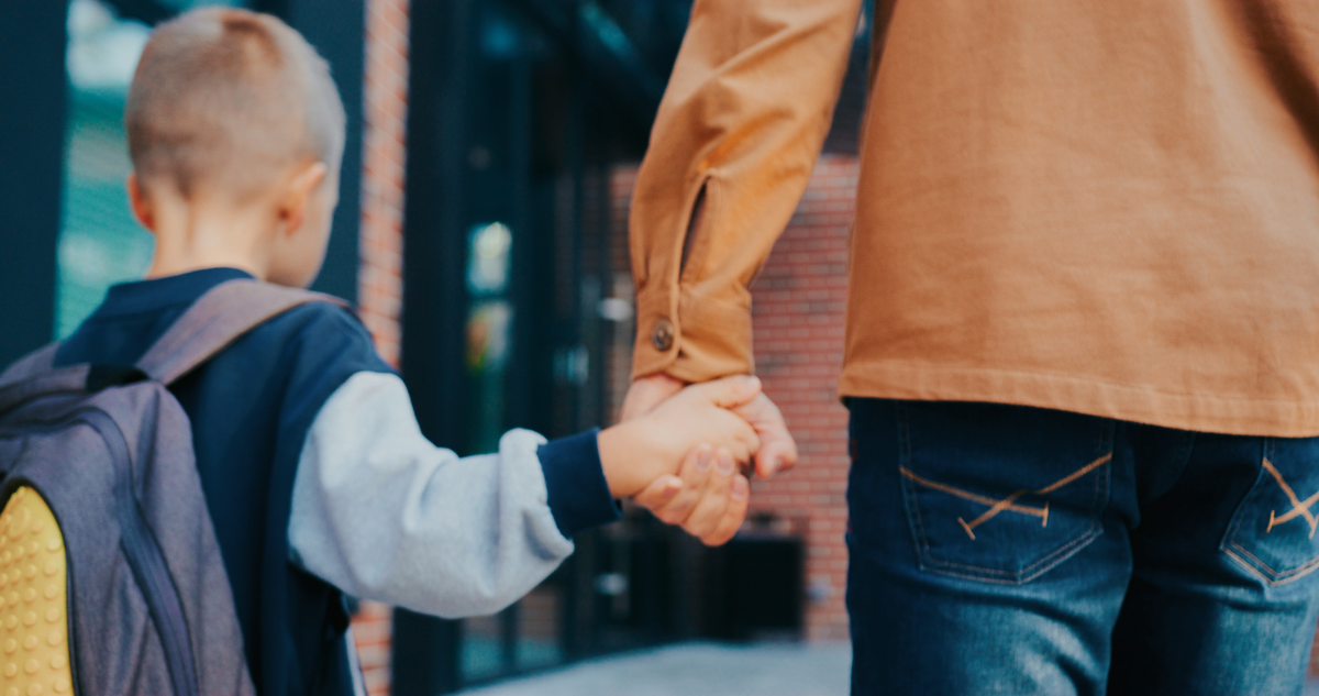 Blick von hinten auf den kleinen kaukasischen Jungen, der mit seinem fürsorglichen Vater zusammen geht. Vater, der den kleinen Jungen in der Schule abgesetzt hat. Er hält seine Hand. Rucksack mit Büchern. Moderne Schule.