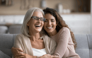 Fröhlich aufgeregt Mutter und erwachsene Tochter Frau, die sich zu Hause amüsiert, mit Kopf berühren, auf dem Sofa sitzen, mit geschlossenen Augen lachen, lustiges Gespräch genießen, Freizeit, Familienaffektion