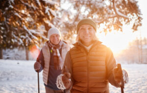 Waist up portrait of smiling mature couple enjoying Nordic walk in winter forest at sunset