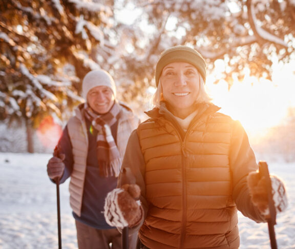 Waist up portrait of smiling mature couple enjoying Nordic walk in winter forest at sunset