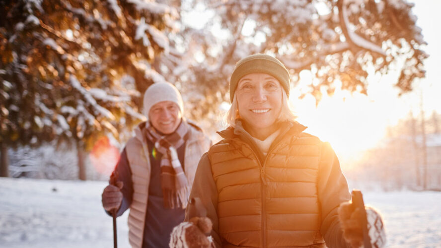 Waist up portrait of smiling mature couple enjoying Nordic walk in winter forest at sunset