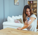 Junge Frau lächelt, während sie mit ihrem Smartphone, um Gesundheitsdaten von einem tragbaren Gerät zu Hause überprüfen. Das Bild zeigt moderne Technologie und Gesundheitsmanagement in einem ungezwungenen Rahmen.