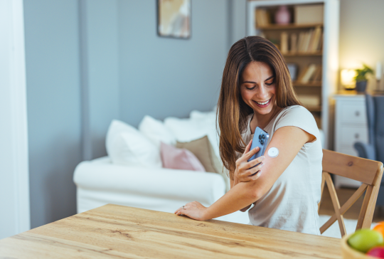 Junge Frau lächelt, während sie mit ihrem Smartphone, um Gesundheitsdaten von einem tragbaren Gerät zu Hause überprüfen. Das Bild zeigt moderne Technologie und Gesundheitsmanagement in einem ungezwungenen Rahmen.