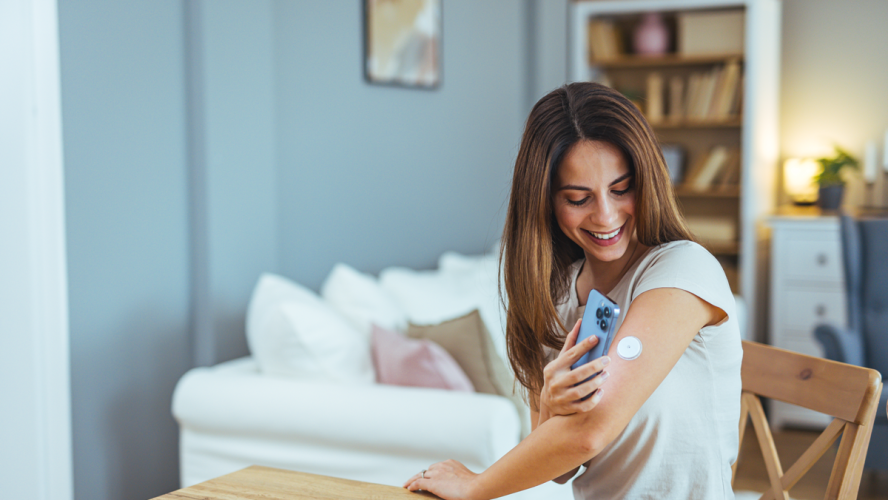 Junge Frau lächelt, während sie mit ihrem Smartphone, um Gesundheitsdaten von einem tragbaren Gerät zu Hause überprüfen. Das Bild zeigt moderne Technologie und Gesundheitsmanagement in einem ungezwungenen Rahmen.