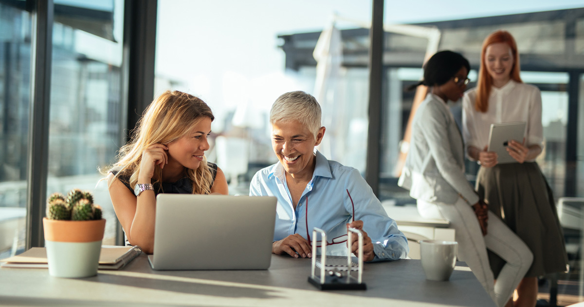Two businesswomen working on a computer. Two women in the background looking at a digital tablet