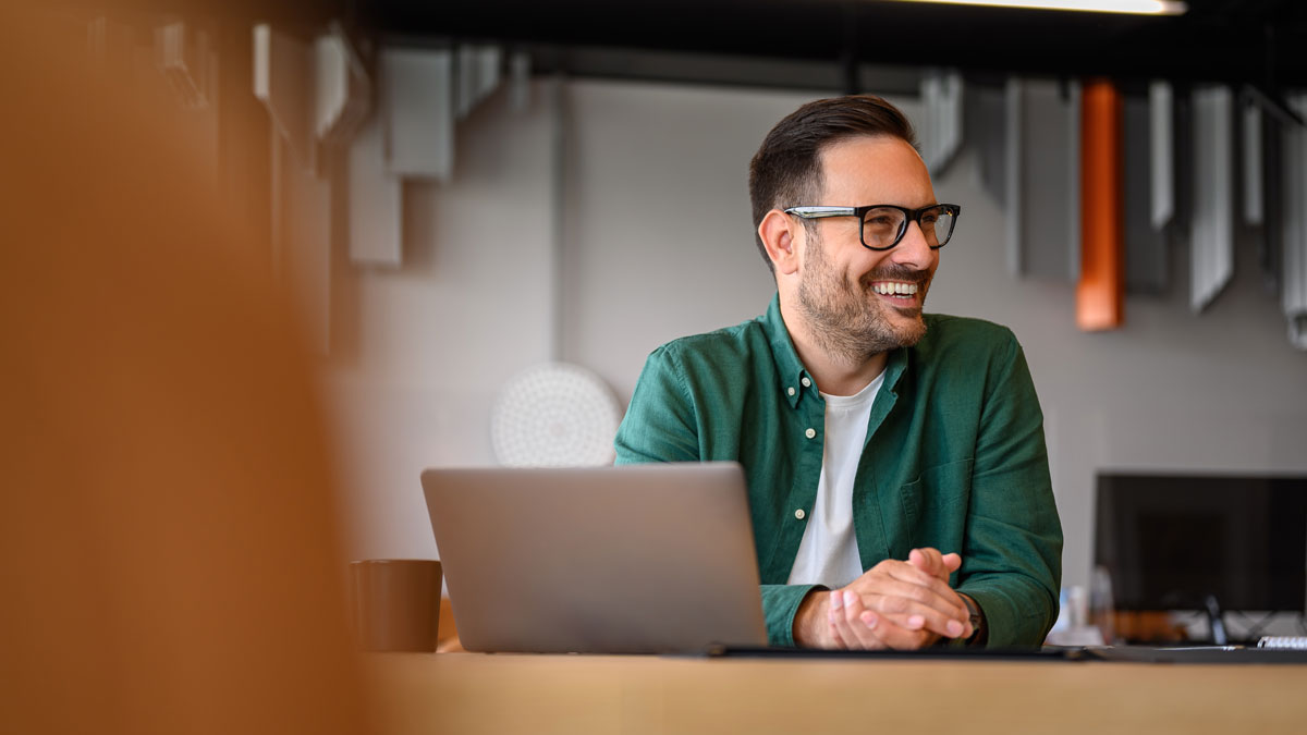 Happy young businessman in eyeglasses looking away while working online over laptop at desk in office